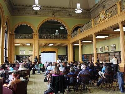 Speakers sitting in front of a presentation screen in a historic building speaking to a full room of audience.