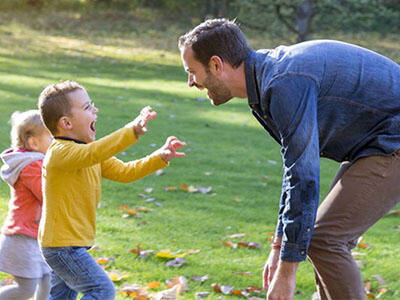 A young boy in a yellow shirt and young girl in a pink jacket are happily running to a crouched man with open arms in a blue shirt.